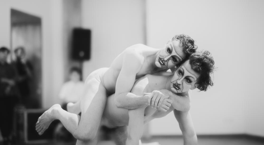 A nude person rests on another nude person's back, the second person is on hands and knees