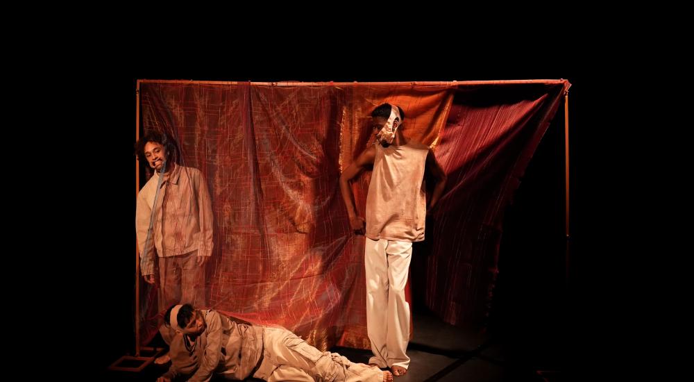 Three performers, two standing, one lying on the floor, in front of a red sheet