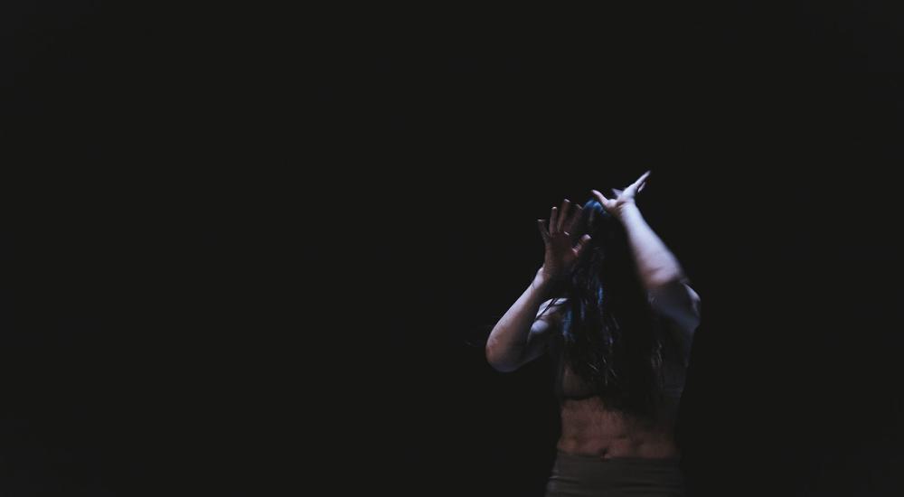 A dancer in front of a dark background holding their arms up in front of their face
