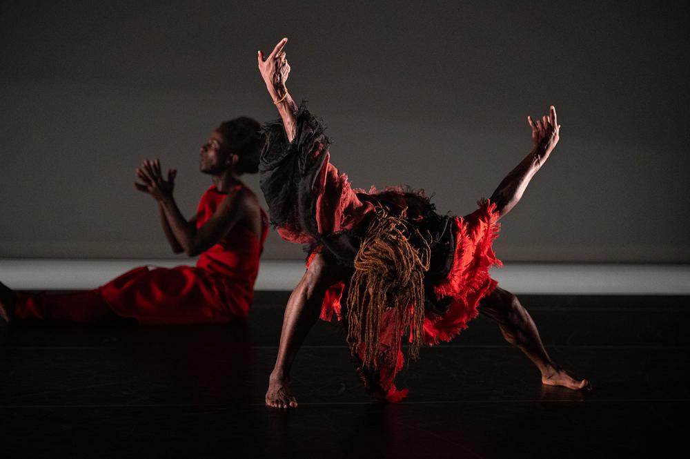 Two dancers, one bending forward, arms outstretched begind him, the other sitting, holding their hands together