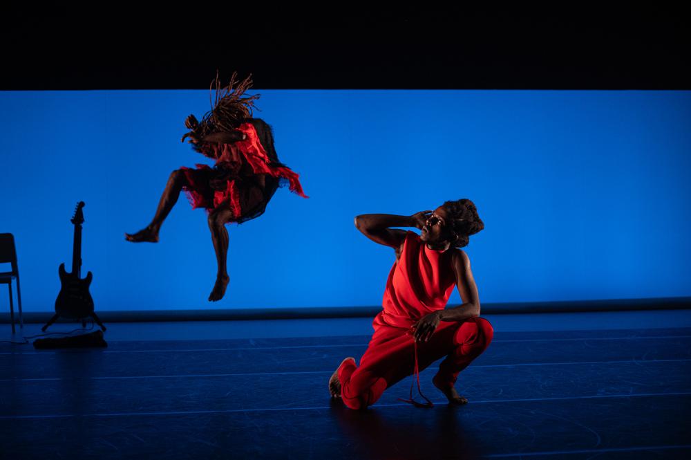 Two Black dancers, one kneeling, one jumping in the air, wearing red in front of a blue screen