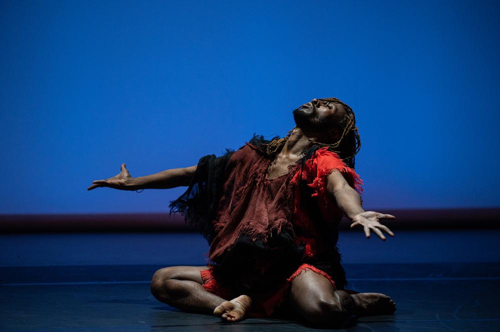 A dancer sitting on the floor, legs to the side, arms outstretched, face aimed towards the sky