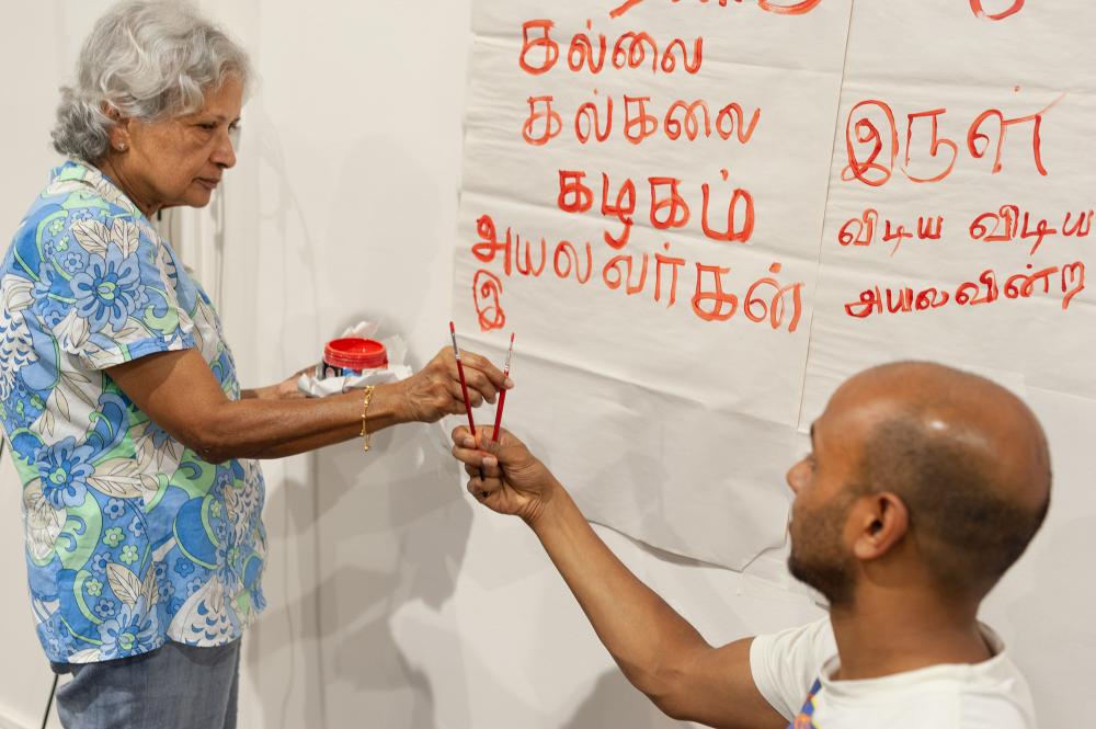 Ahilan handing his mum a paintbrush in front of papers stuck to the wall painted with Tamil script