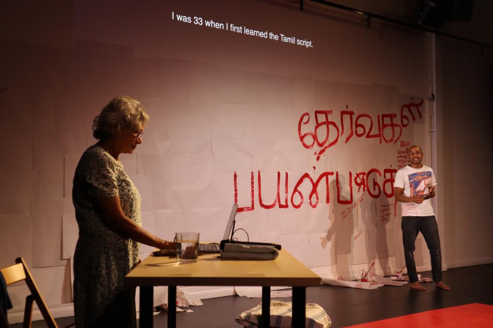 Ahilan standing in front of a screen with Tamil text on it and English surtitles operated by his mum behind a laptop on the side