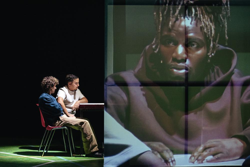 Two actors sitting at a table while a third is projected on a screen