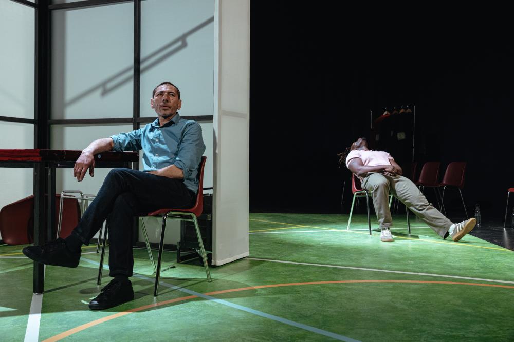 One actor sitting in a chair, while another is lying back in his behind him