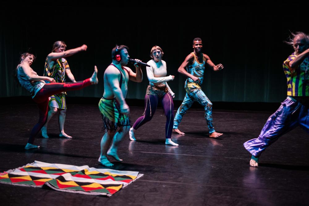 6 dancers wearing colourful outfits, one holding a mic and wearing headphones