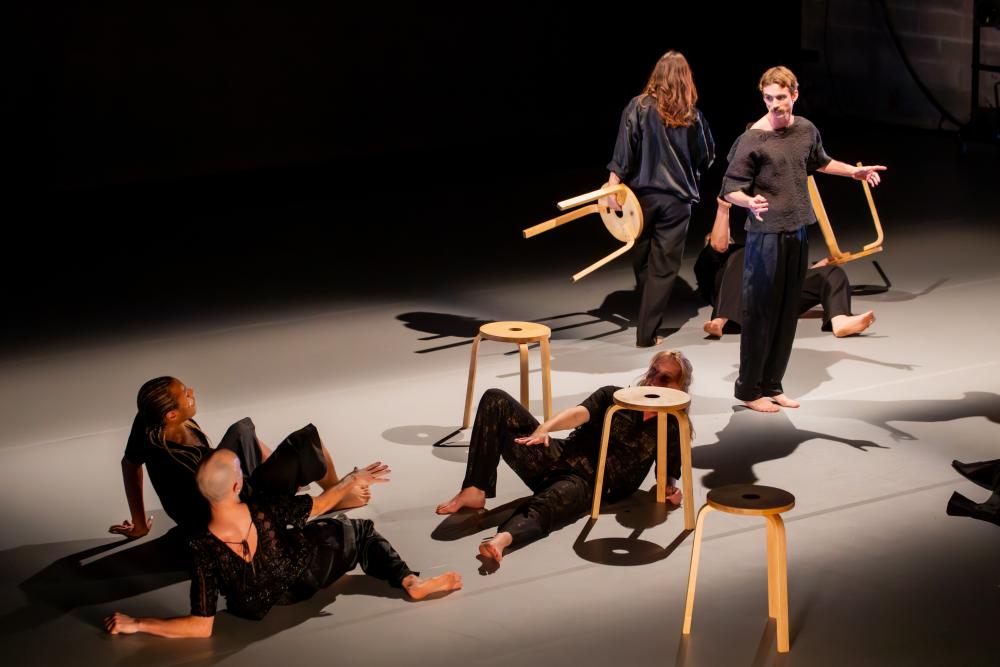 two people standing, carrying stools, while three others are lying on the floor