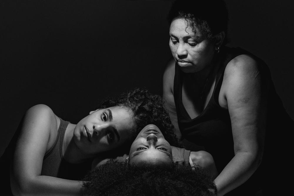 A black-and-white photo of three women holding each other, one sitting, one lying on her back with the last one resting her head on her