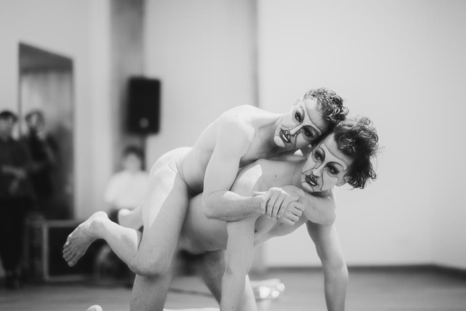 A nude person rests on another nude person's back, the second person is on hands and knees