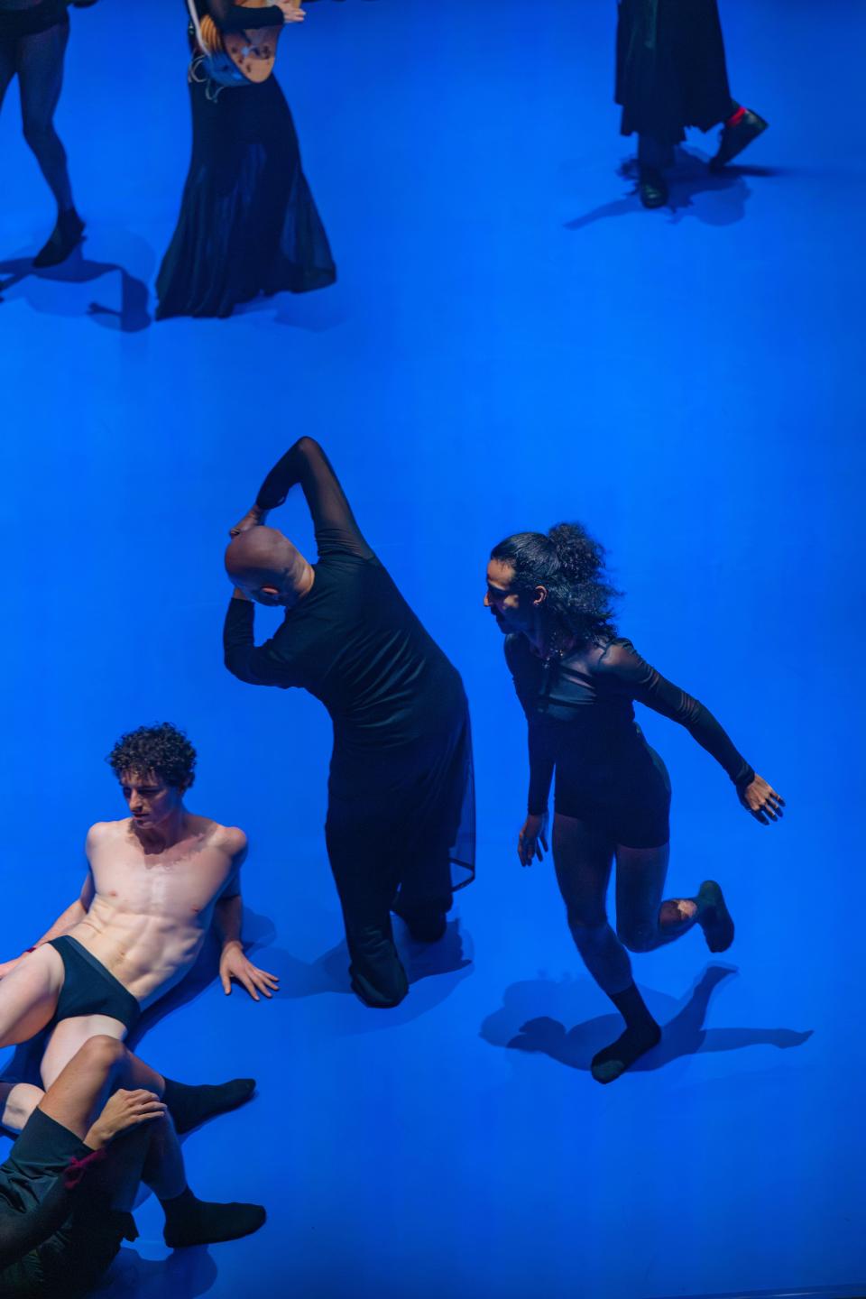 6 performers with black clothes and one male performer only wearing black underwear. All dancing on a bright blue floor