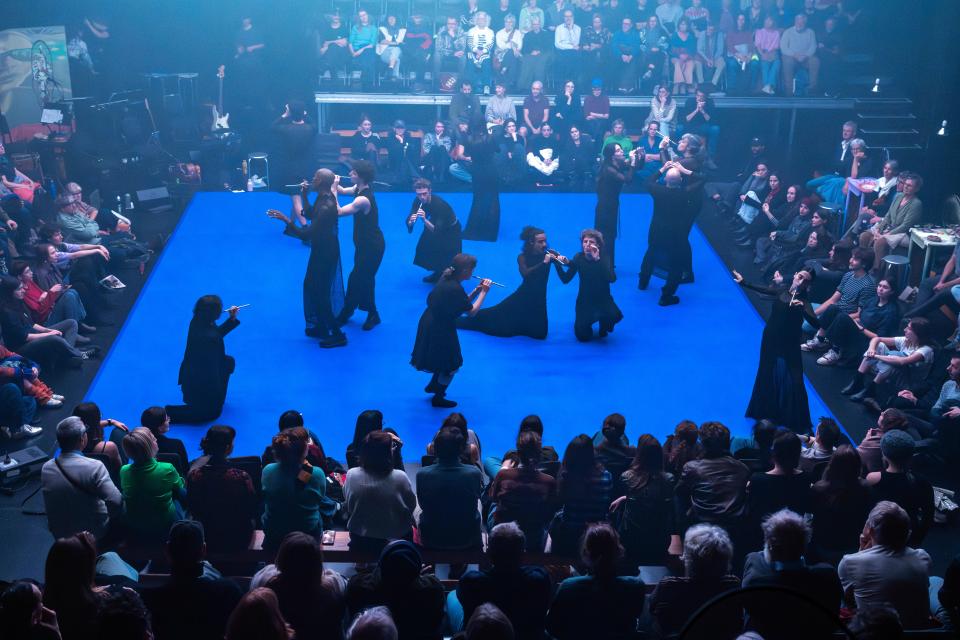 square stage with a bright blue floor, and the audience sitting around it. Different performers on the stage dancing.